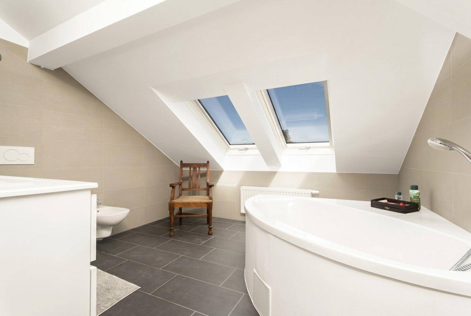 Geräumiges Badezimmer im Dachgeschoss mit Badewanne, Dachfenstern und dunklen Bodenfliesen.