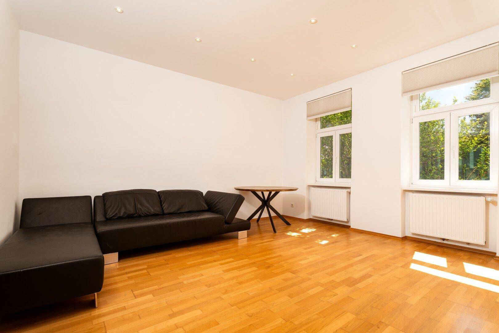 Helles Wohnzimmer mit Parkettboden, einem schwarzen Sofa und zwei großen Fenstern.