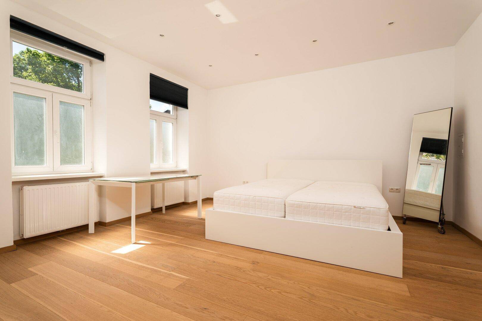 Geräumiges Schlafzimmer mit hellem Parkettboden, einem weißen Bett und viel Tageslicht.