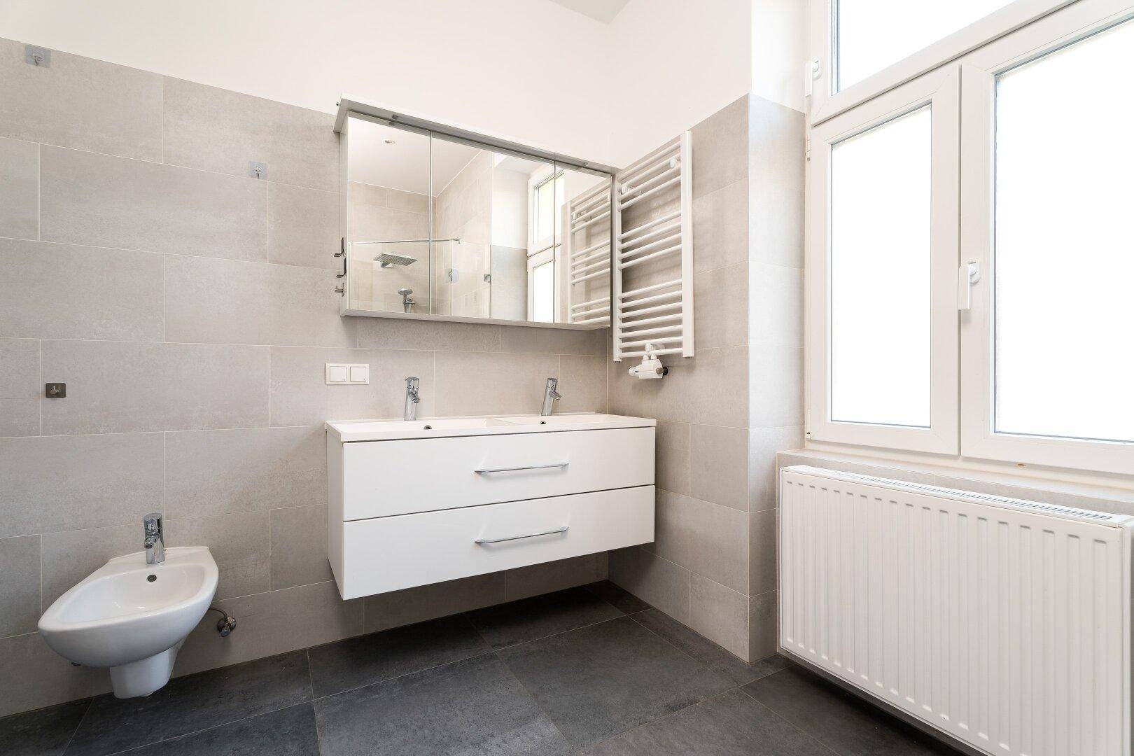 Helles Badezimmer mit Doppelwaschbecken, Bidet, Spiegelschrank und großem Fenster.