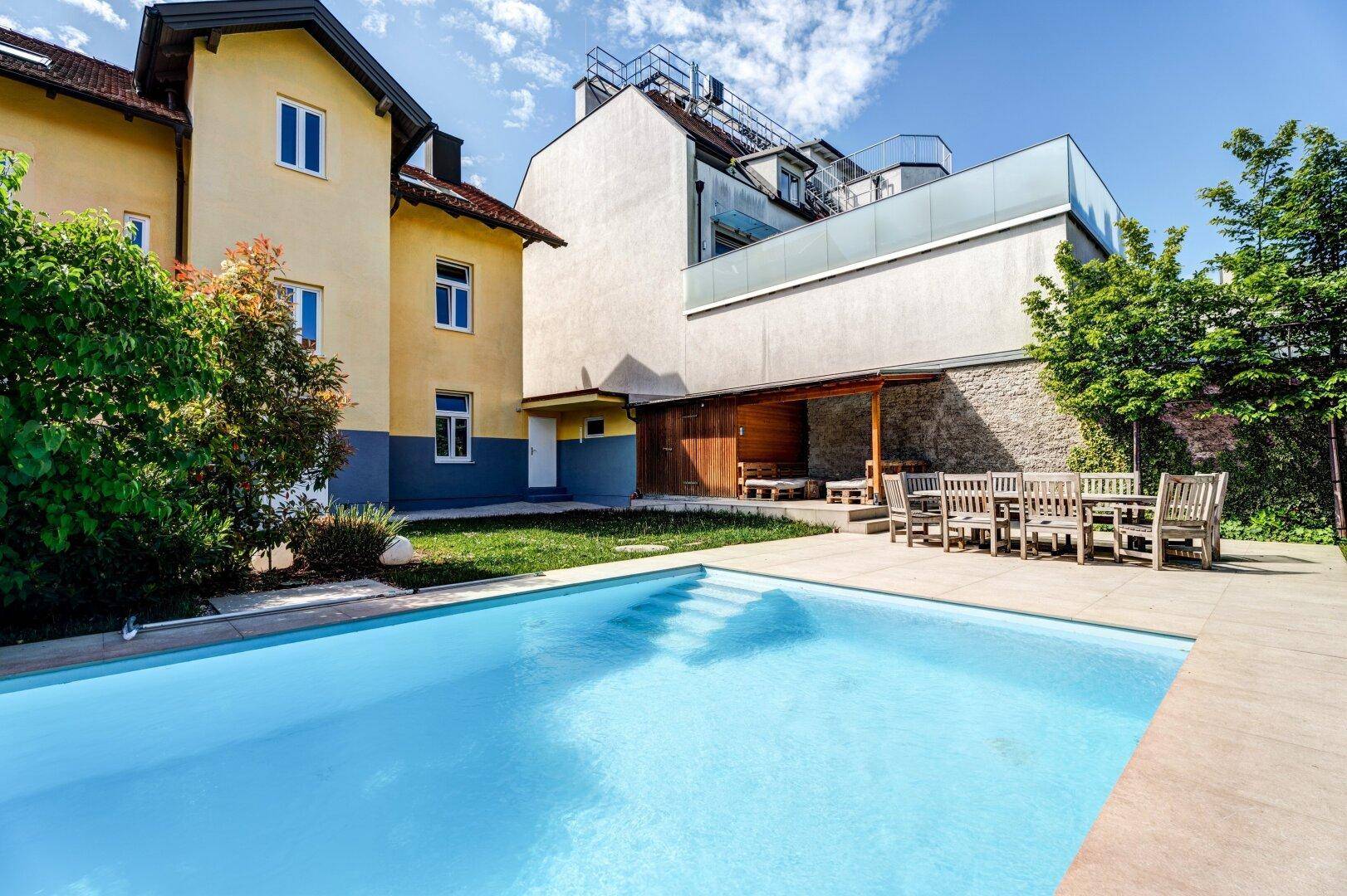 Blick auf den Swimmingpool und den Essbereich im Freien, umgeben von Bäumen und Gebäuden.