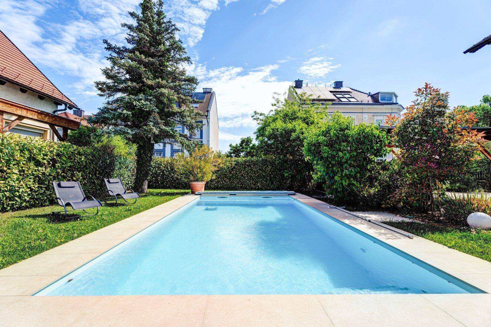 Langer Swimmingpool im Garten mit Liegestühlen und üppiger Bepflanzung unter blauem Himmel.