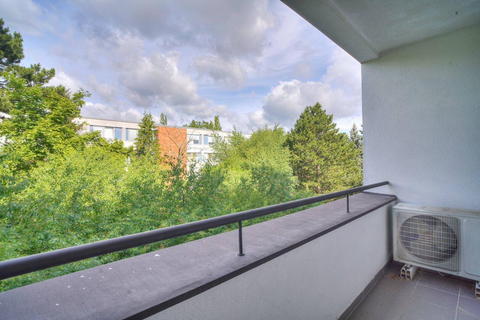 Balkon mit Klimaanlage und weitem Blick auf Bäume und umliegende Gebäude.