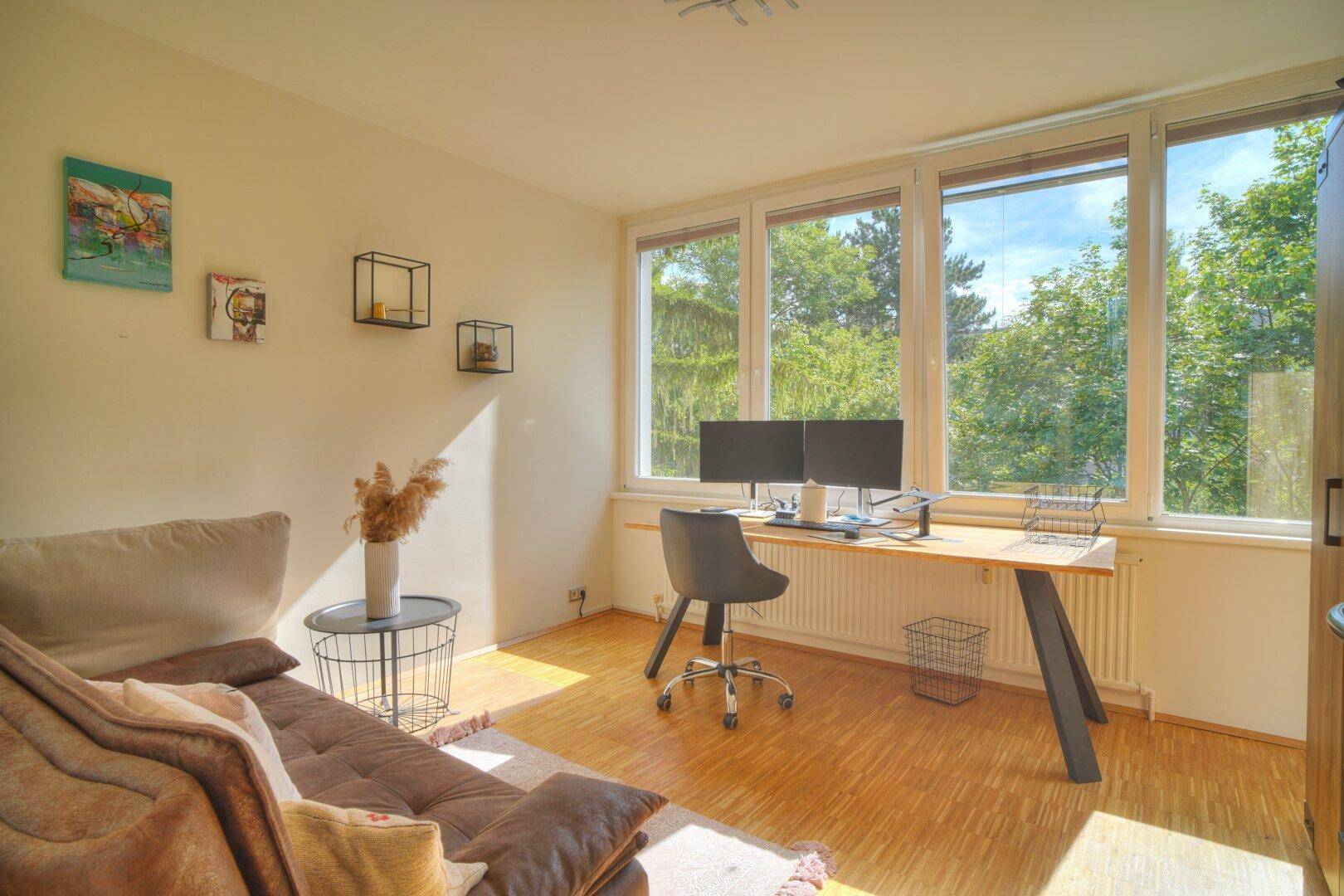 Helles Arbeitszimmer mit Schreibtisch, Sofa und großen Fenstern mit Blick ins Grüne.