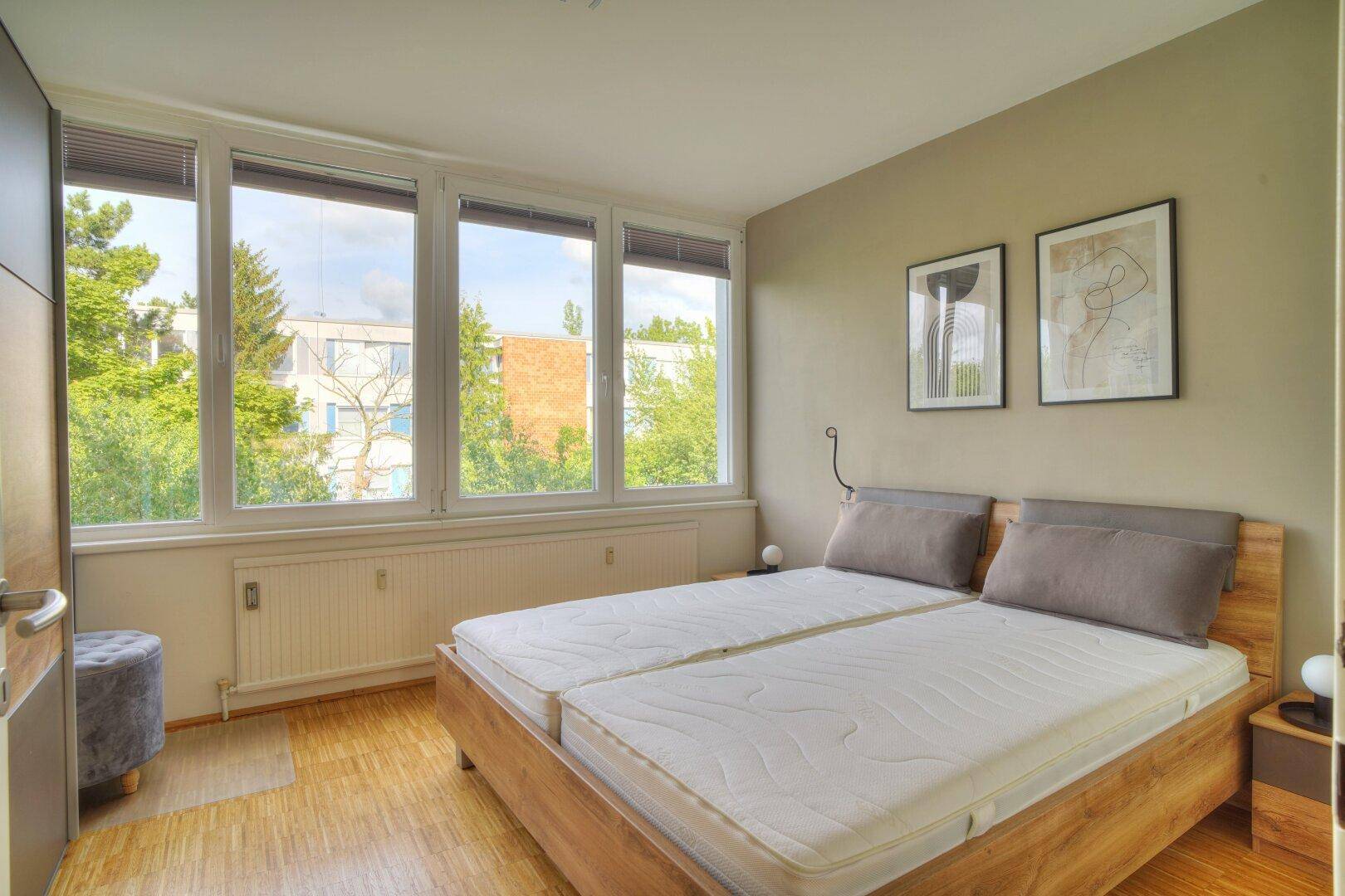 Helles Schlafzimmer mit Doppelbett, Holzfußboden und großen Fenstern mit Blick ins Grüne.