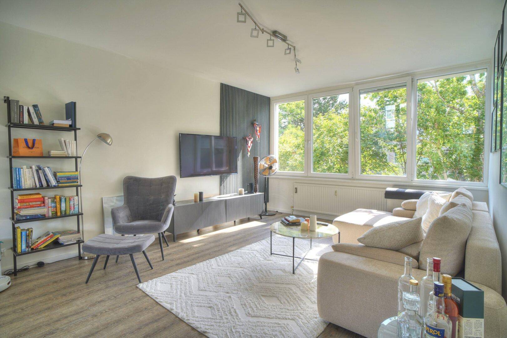 Modern eingerichtetes Wohnzimmer mit beigem Sofa, grauem Sessel und großen Fenstern.