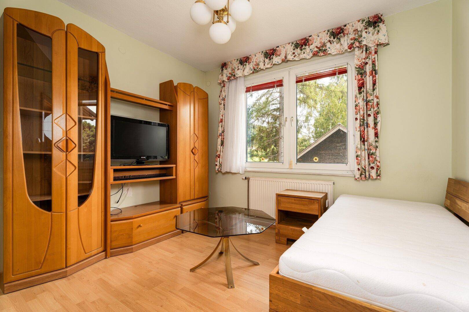 Helles Schlafzimmer mit Einzelbett, Holzmöbeln und Blick ins Grüne.
