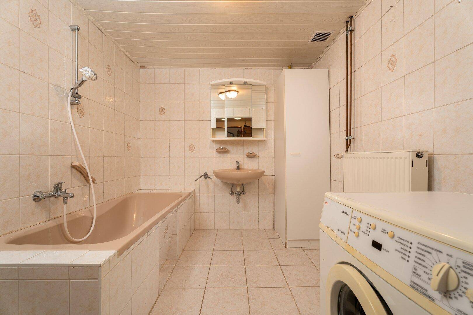 Badezimmer mit Badewanne, Waschbecken, Spiegel und Waschmaschine, ausgestattet mit beigen Fliesen.