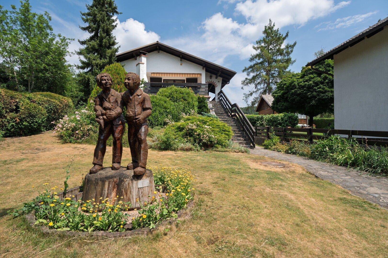 Idyllischer Gartenbereich vor dem Haus mit Rasenfläche, Steinweg und einer charmanten Holzskulptur.