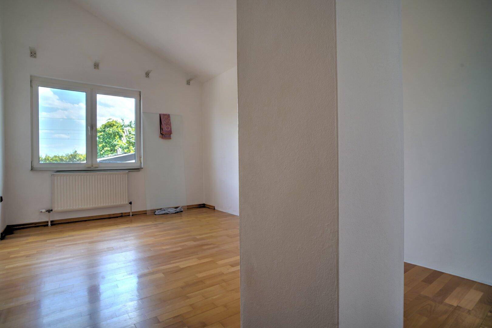 Geräumiges, helles Zimmer mit Holzboden, weißen Wänden und großem Fenster.