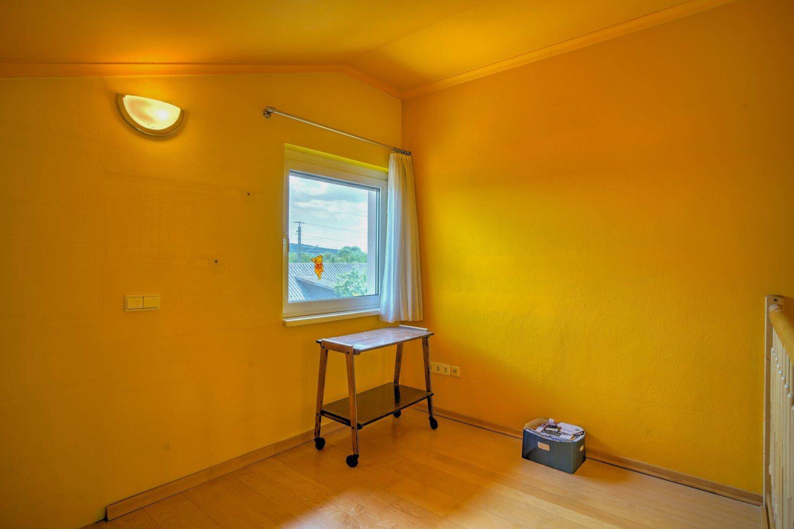 Sonniger Raum mit gelben Wänden, Fenster und Holzboden, vielseitig nutzbar.