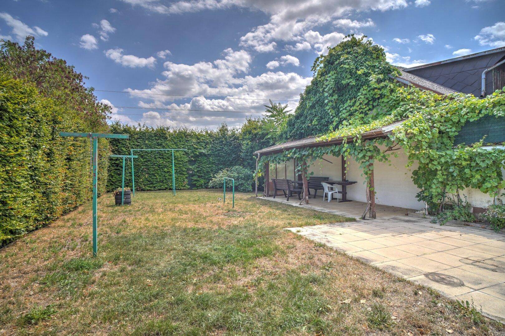 Großer Garten mit Rasenfläche, überdachter Terrasse und Hecke.