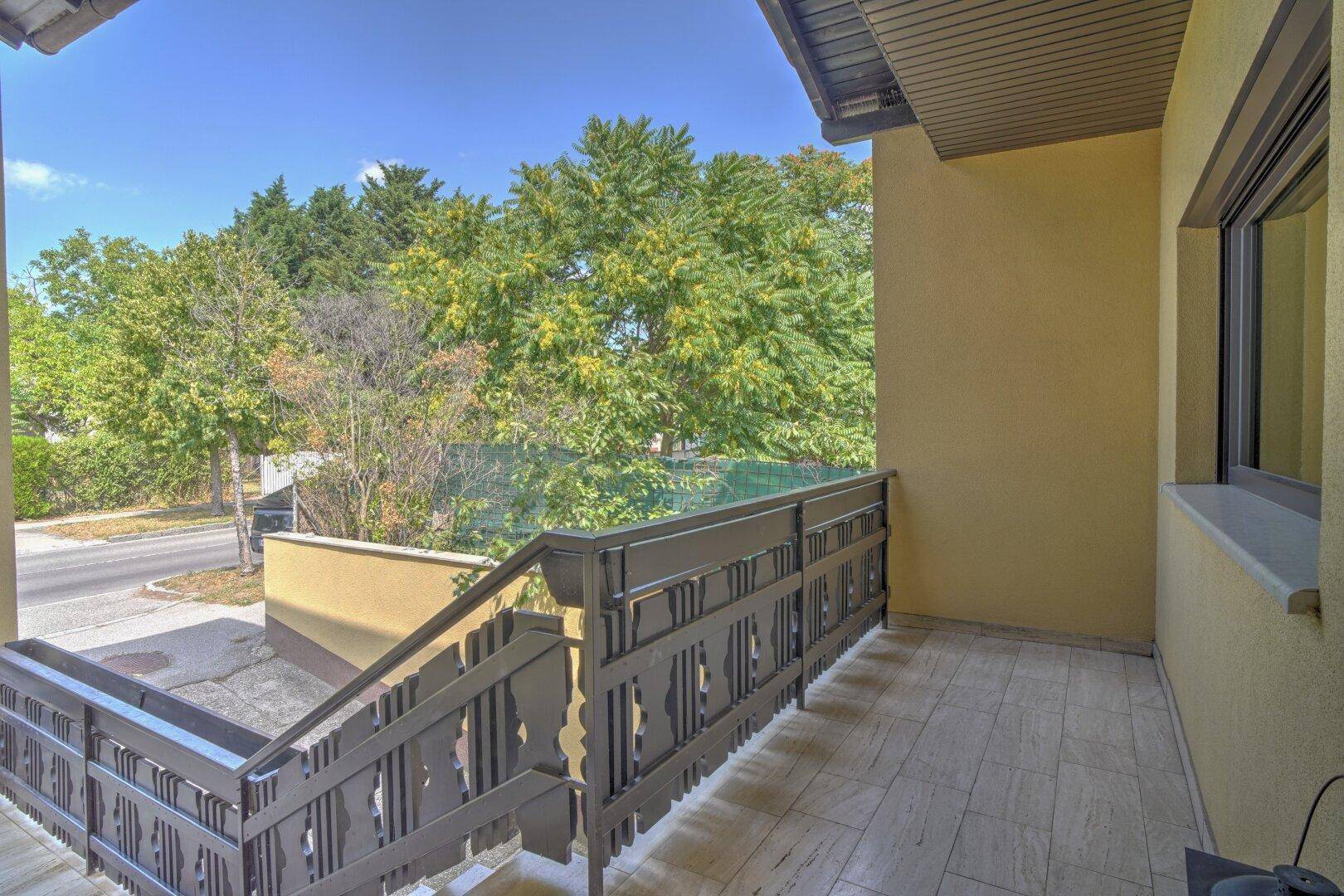 Balkon mit Fliesenboden, Geländer und Blick auf die Straße und Bäume.