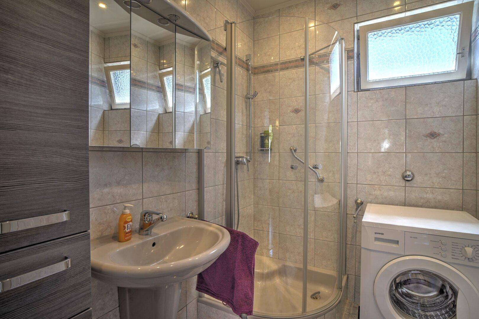 Kleines Badezimmer mit Duschkabine, Waschbecken und Waschmaschine, hell gefliest.