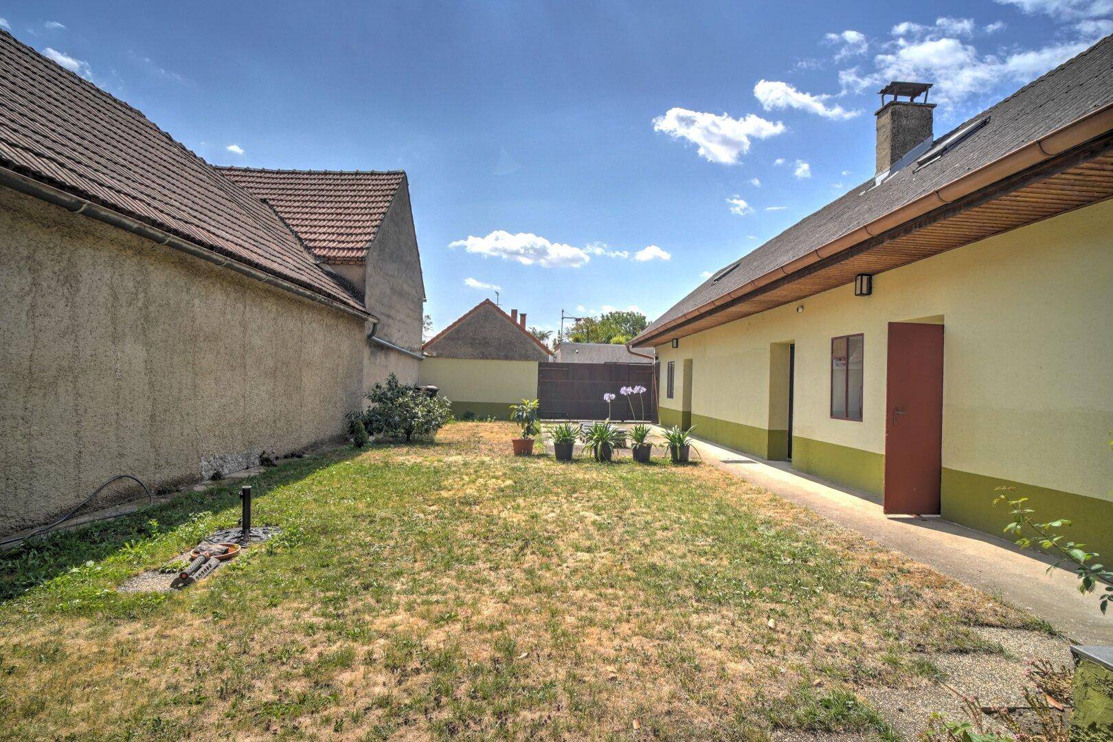 Kleiner Gartenbereich mit Rasen und Topfpflanzen, umgeben von Gebäuden unter blauem Himmel.