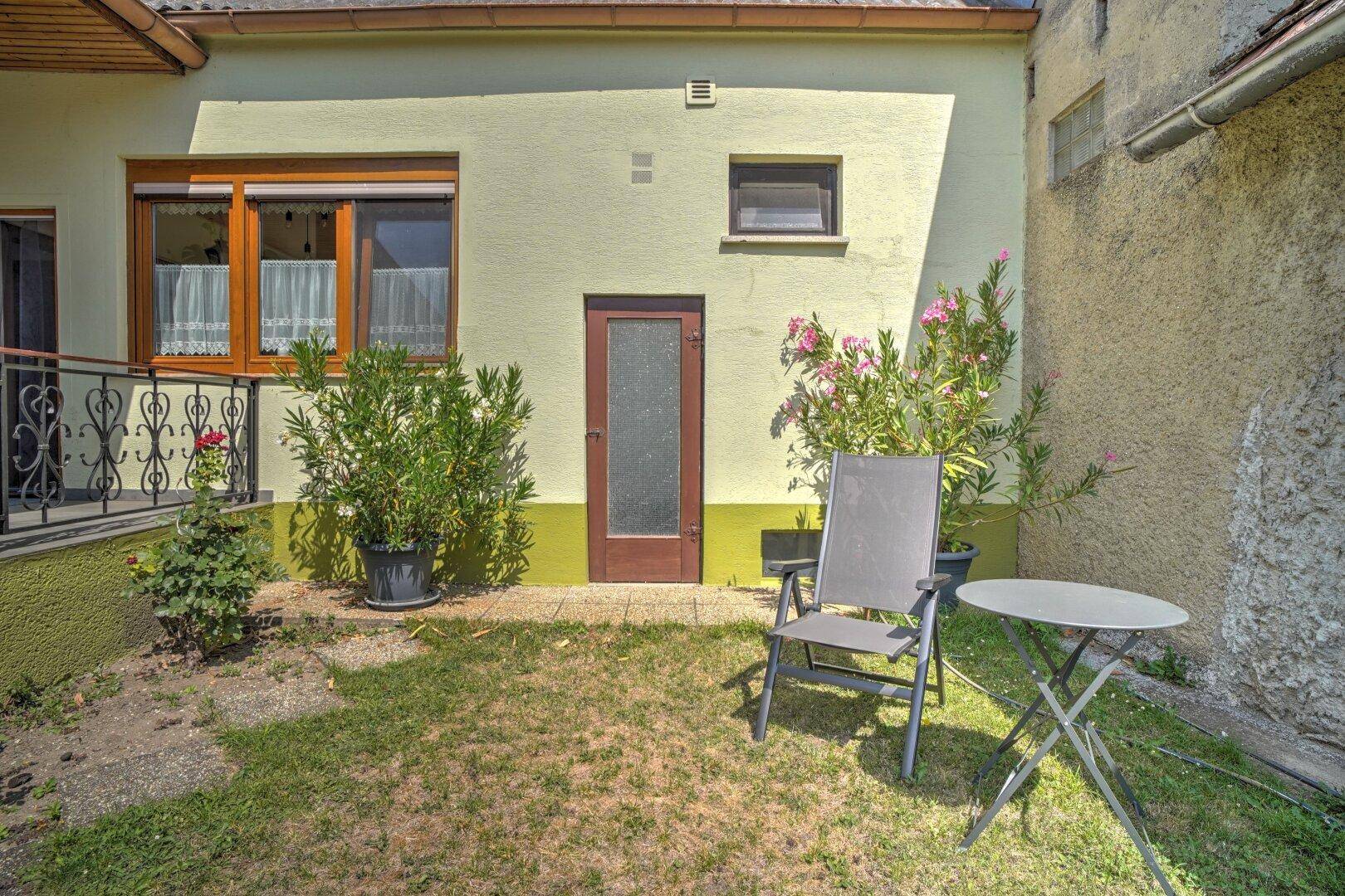 Kleiner Gartenbereich mit Rasen und Sitzgelegenheiten, umgeben von blühenden Pflanzen.