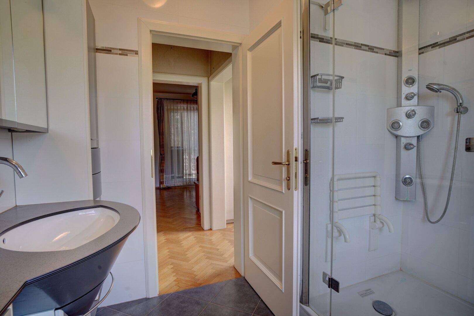 Badezimmer mit moderner Dusche, Waschbecken und Zugang zu einem weiteren Raum.