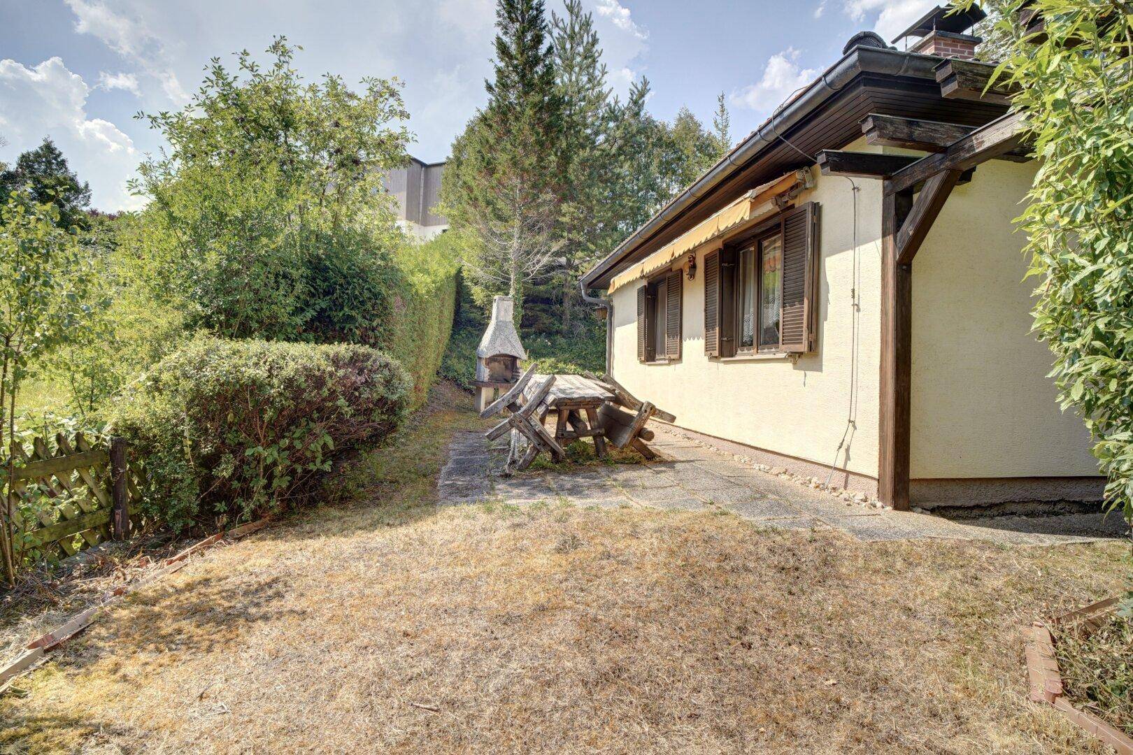 Gartenbereich mit Grillstelle und rustikalem Holztisch, umgeben von Hecken und Bäumen.