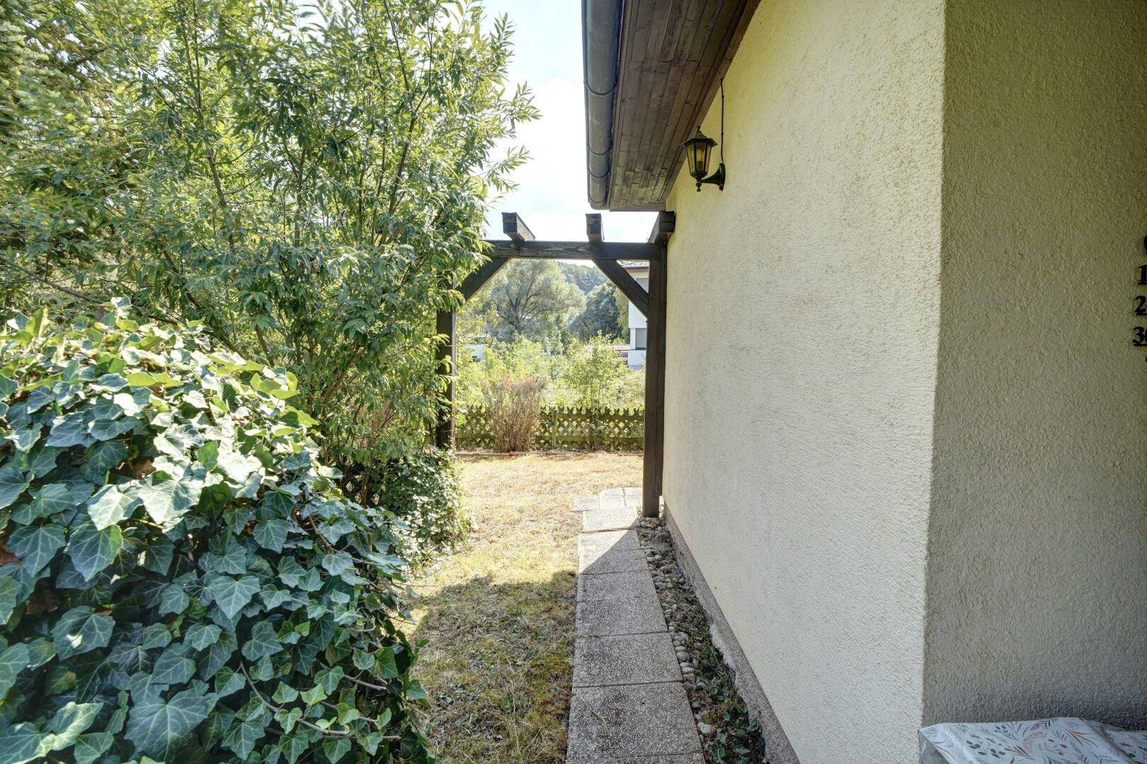Schmaler Gartenweg entlang der Hauswand, der zu einem hölzernen Torbogen und weiteren Grünflächen führt.