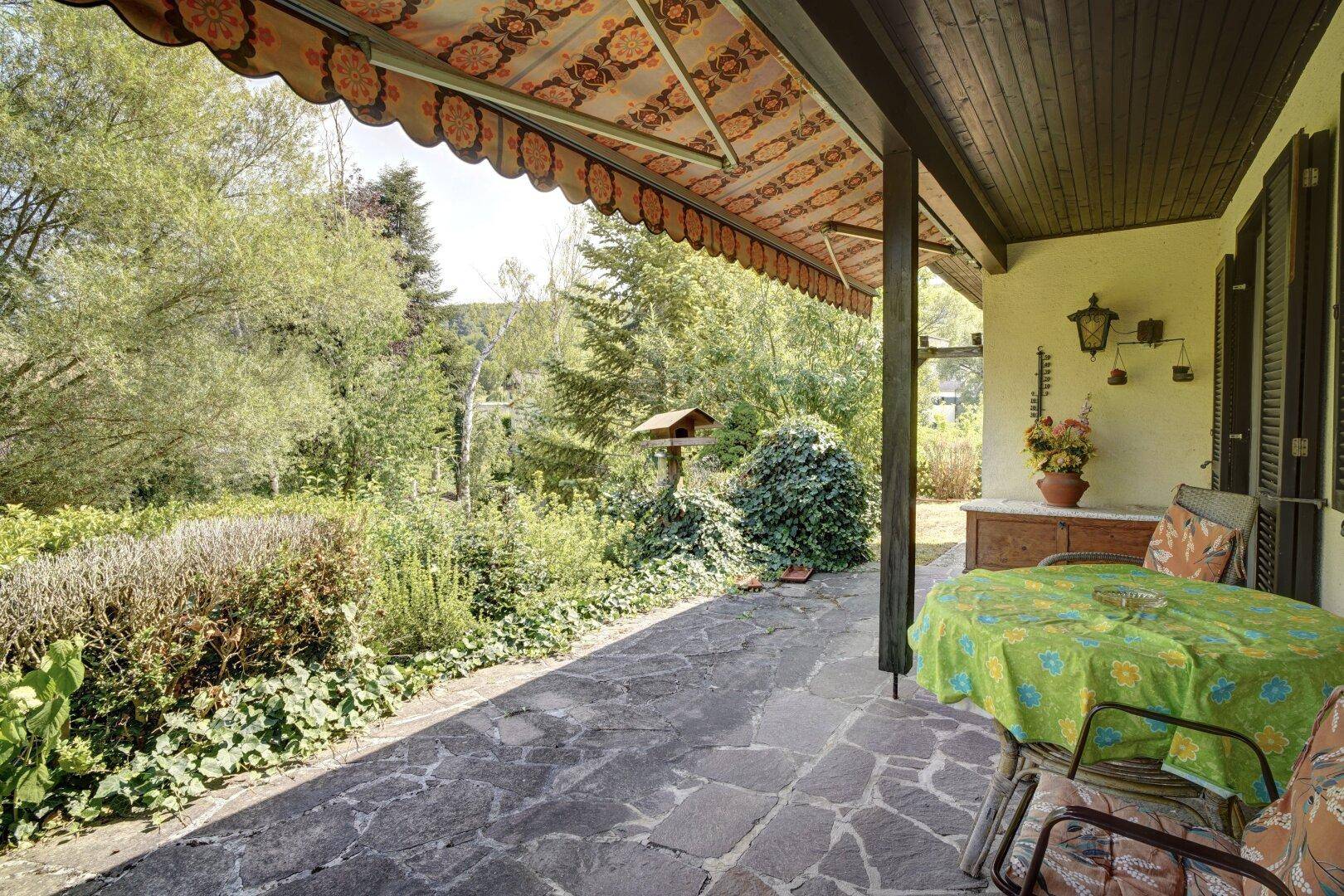 Gepflasterte Terrasse mit Markise, Gartentisch und Stühlen, umgeben von Pflanzen.