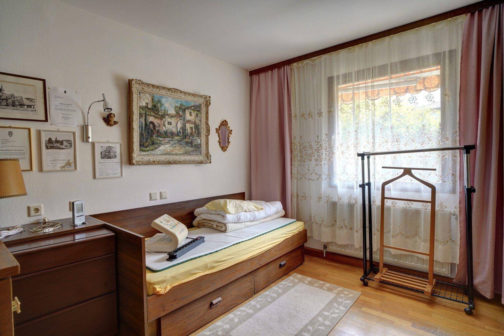 Helles Schlafzimmer mit Einzelbett, Holzkommode und Fenster mit Vorhängen.