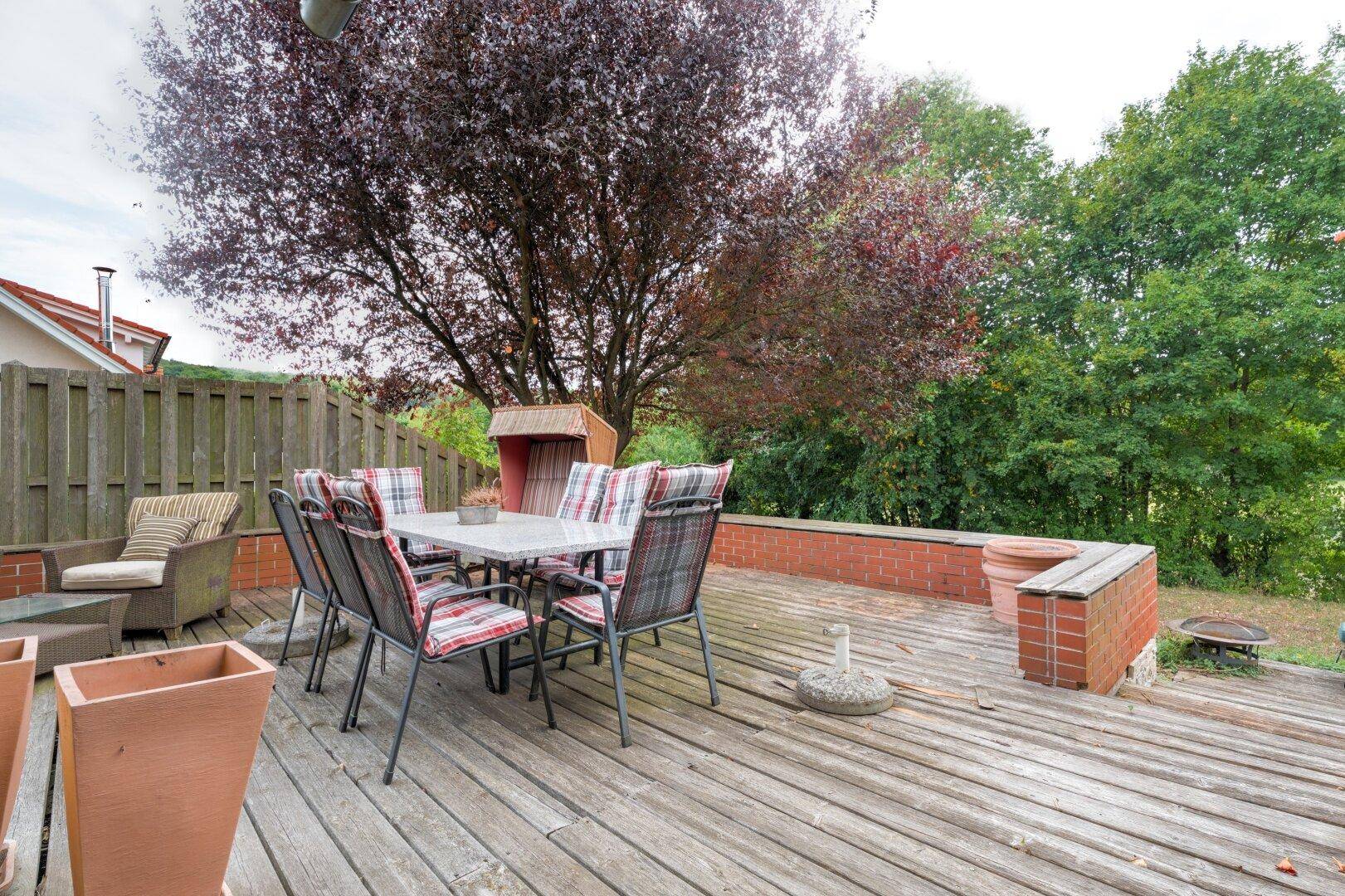 Große Holzterrasse mit Essbereich und Blick auf den grünen Garten und alte Bäume.