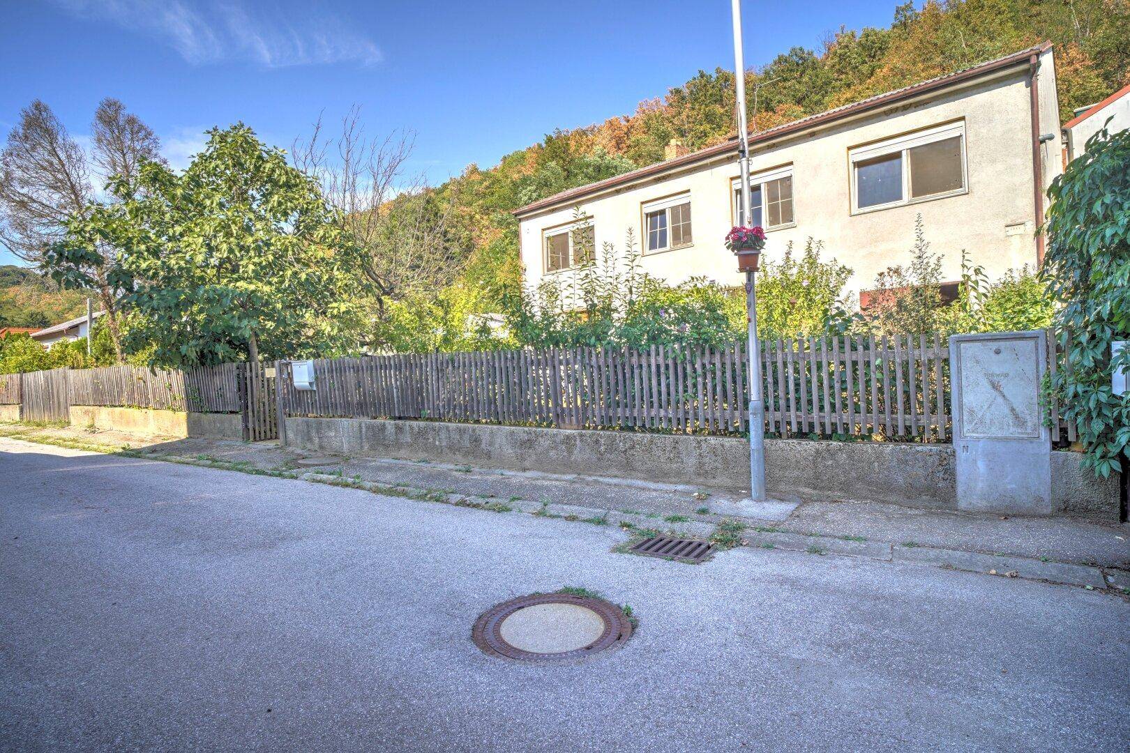 Blick von der Straße auf das Haus mit Holzzaun und bewaldeten Hügeln im Hintergrund.