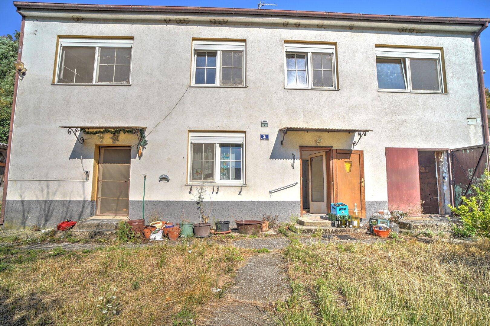 Vorderansicht des Hauses mit zwei Eingängen, mehreren Fenstern und einem ungepflegten Vorgarten.