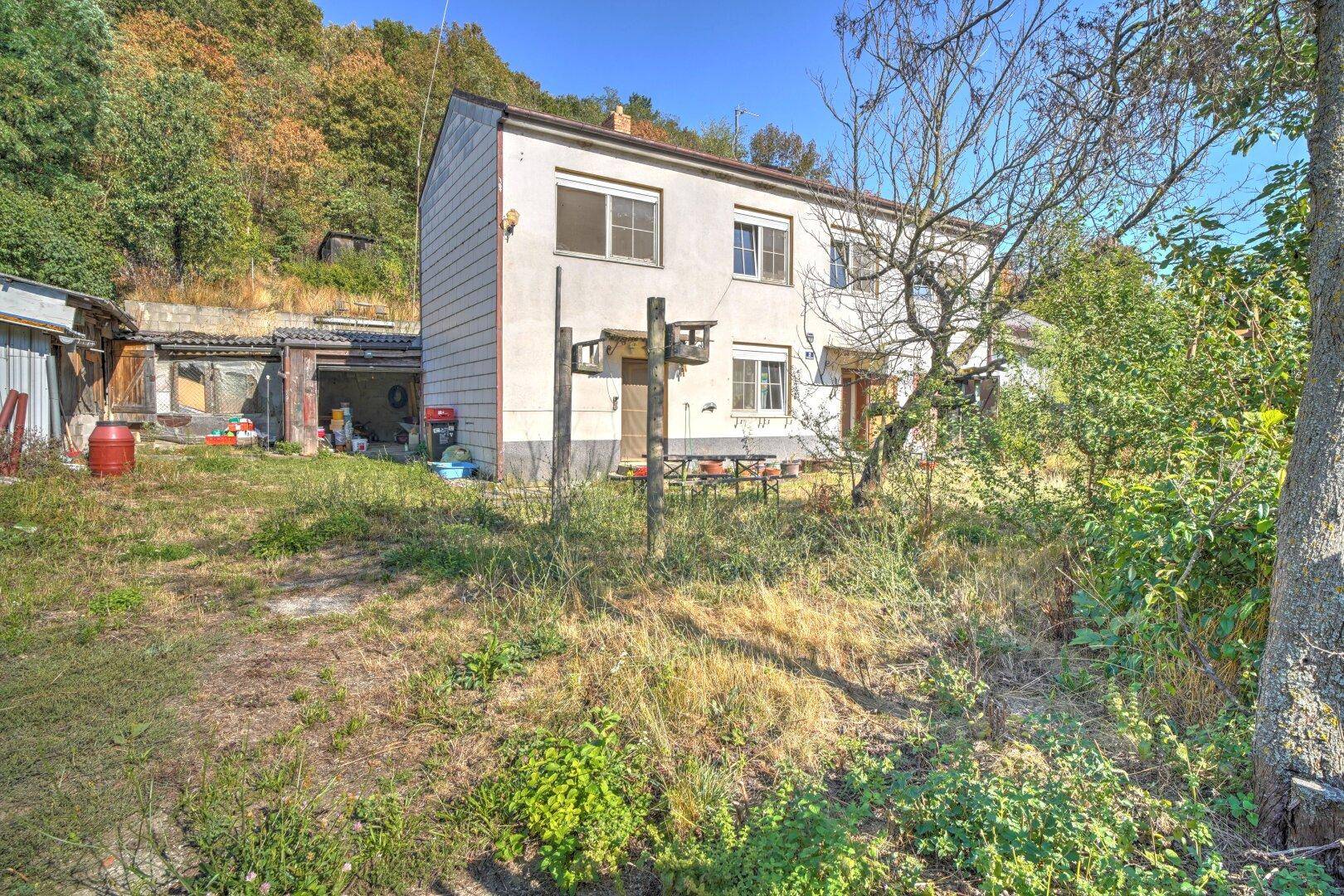 Rückansicht des Hauses mit weitläufigem Garten, Nebengebäuden und bewaldetem Hang im Hintergrund.
