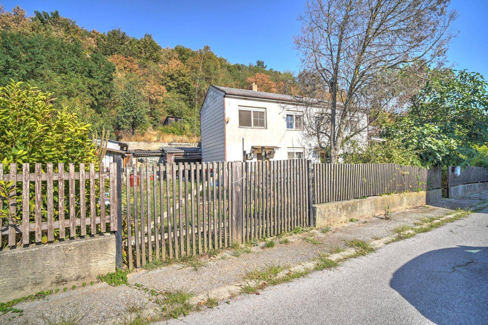 Blick von der Straße auf das Grundstück mit Holzzaun und dem Haus im Hintergrund, umgeben von Natur.