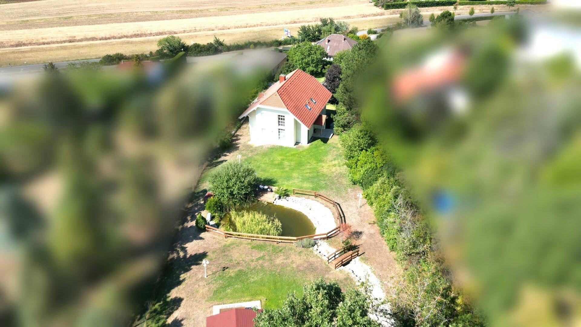 Luftaufnahme des Hauses mit rotem Dach, umgeben von einem Teich, Garten und weiten Feldern.