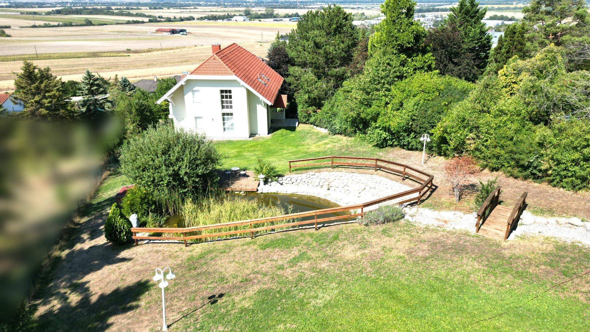 Luftaufnahme des Hauses mit großem Garten, Teich und Holzbrücke, umgeben von viel Grün.