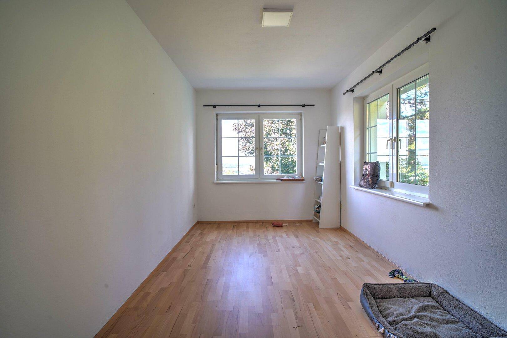 Helles, leeres Zimmer mit Holzboden und zwei Fenstern, bereit zur individuellen Gestaltung.