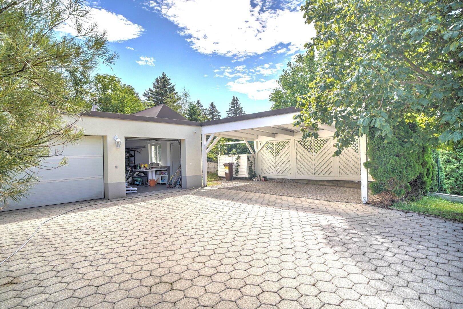 Ansicht der Garage und des Carports mit breiter Auffahrt, umgeben von Bäumen und Sträuchern.