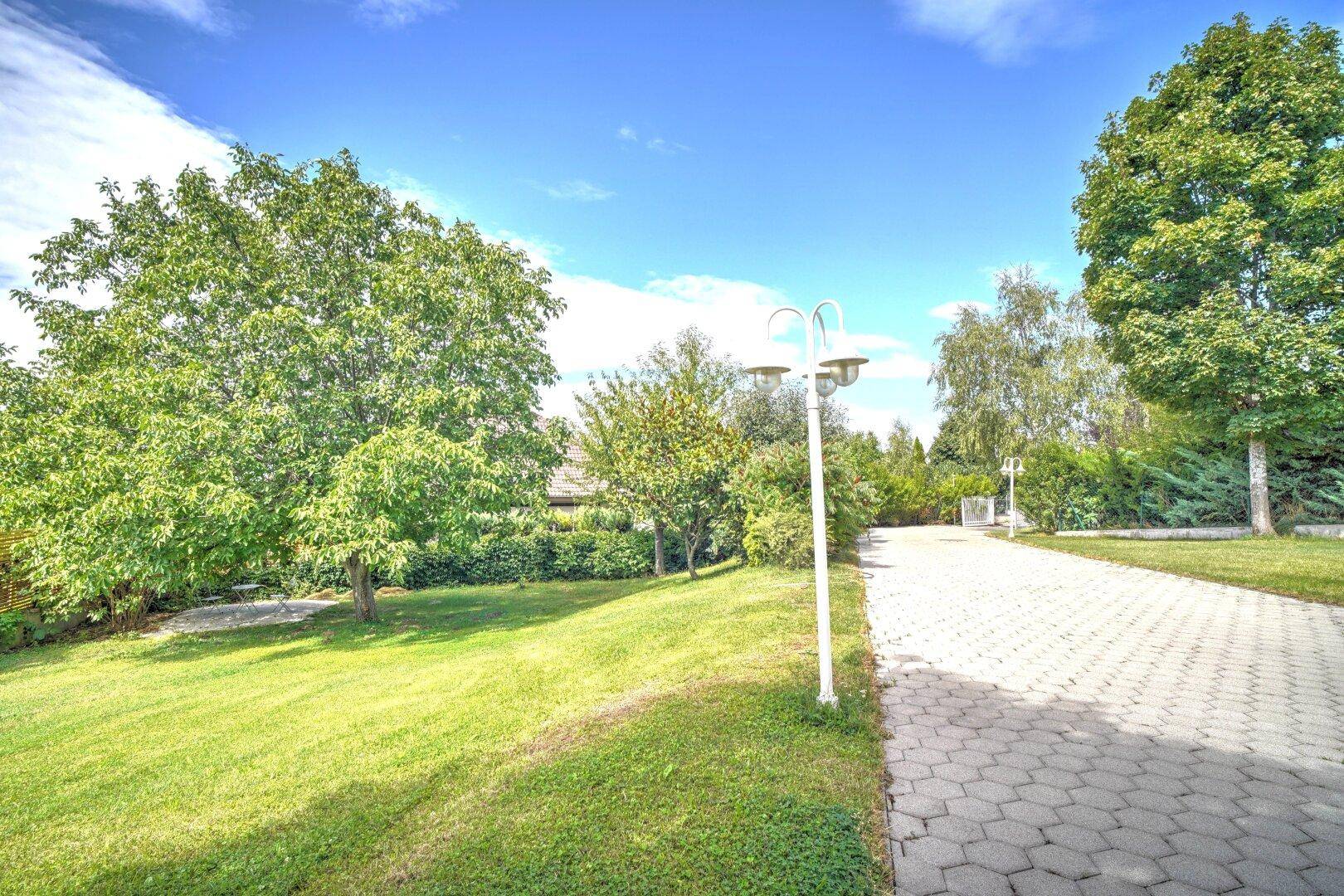 Gepflasterter Weg führt durch den gepflegten Garten mit Rasenflächen und Bäumen unter blauem Himmel.