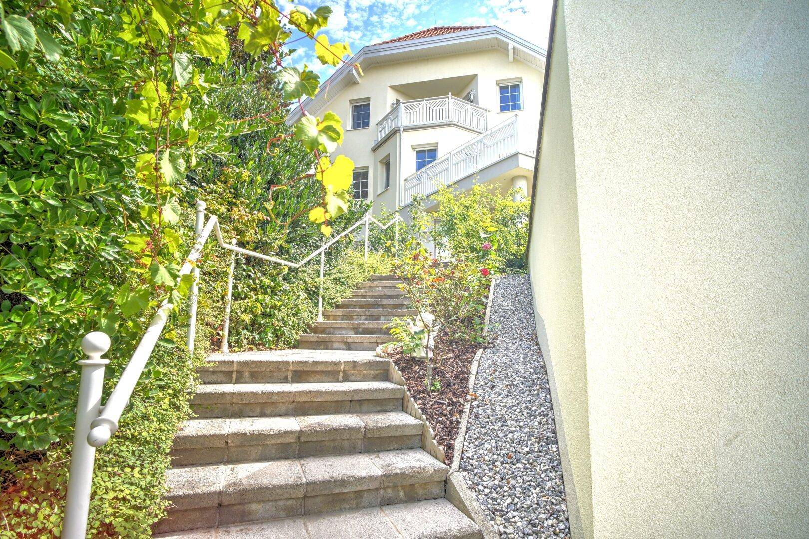 Elegante Steintreppe mit weißem Geländer führt durch üppiges Grün zum Haupthaus hinauf.