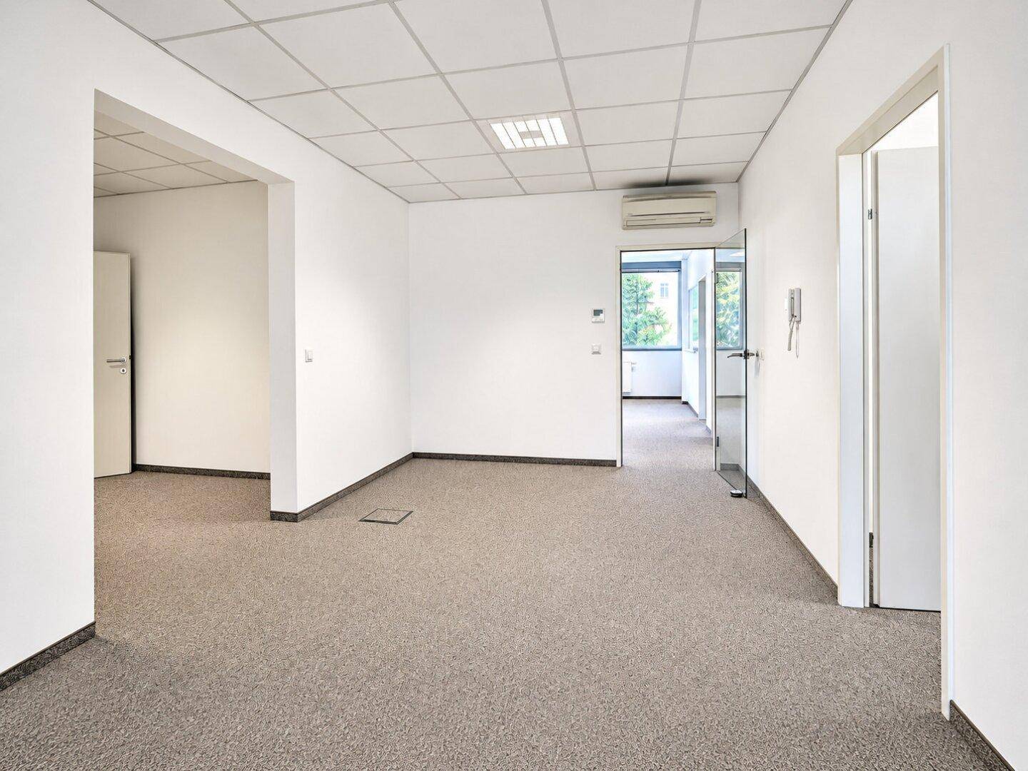 Leerer Bürobereich mit Teppichboden, Klimaanlage und Zugang zu weiteren Räumen.