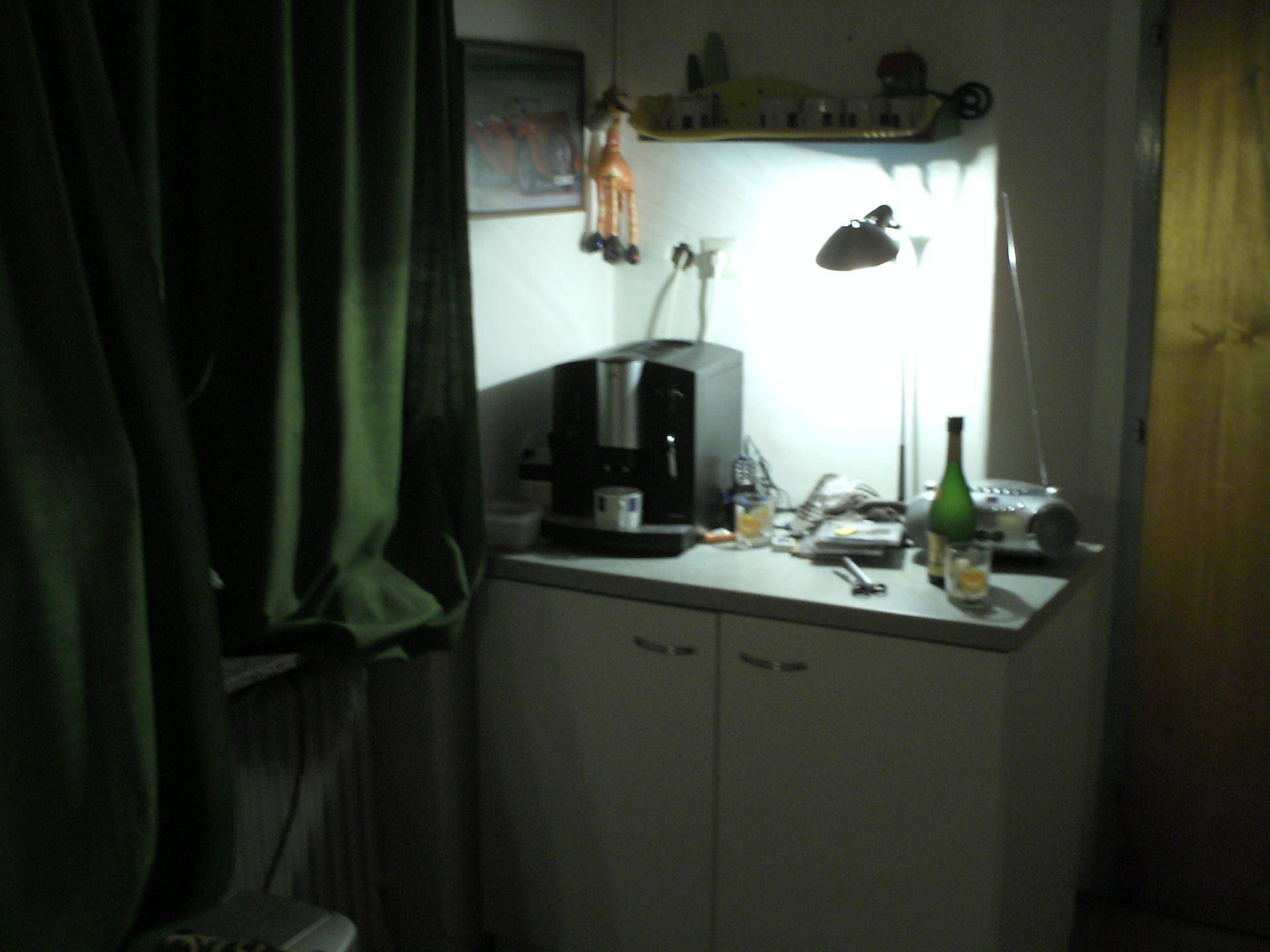 Kleine Küchenzeile mit Kaffeemaschine, Flaschen und Schränken unter einem Fenster.