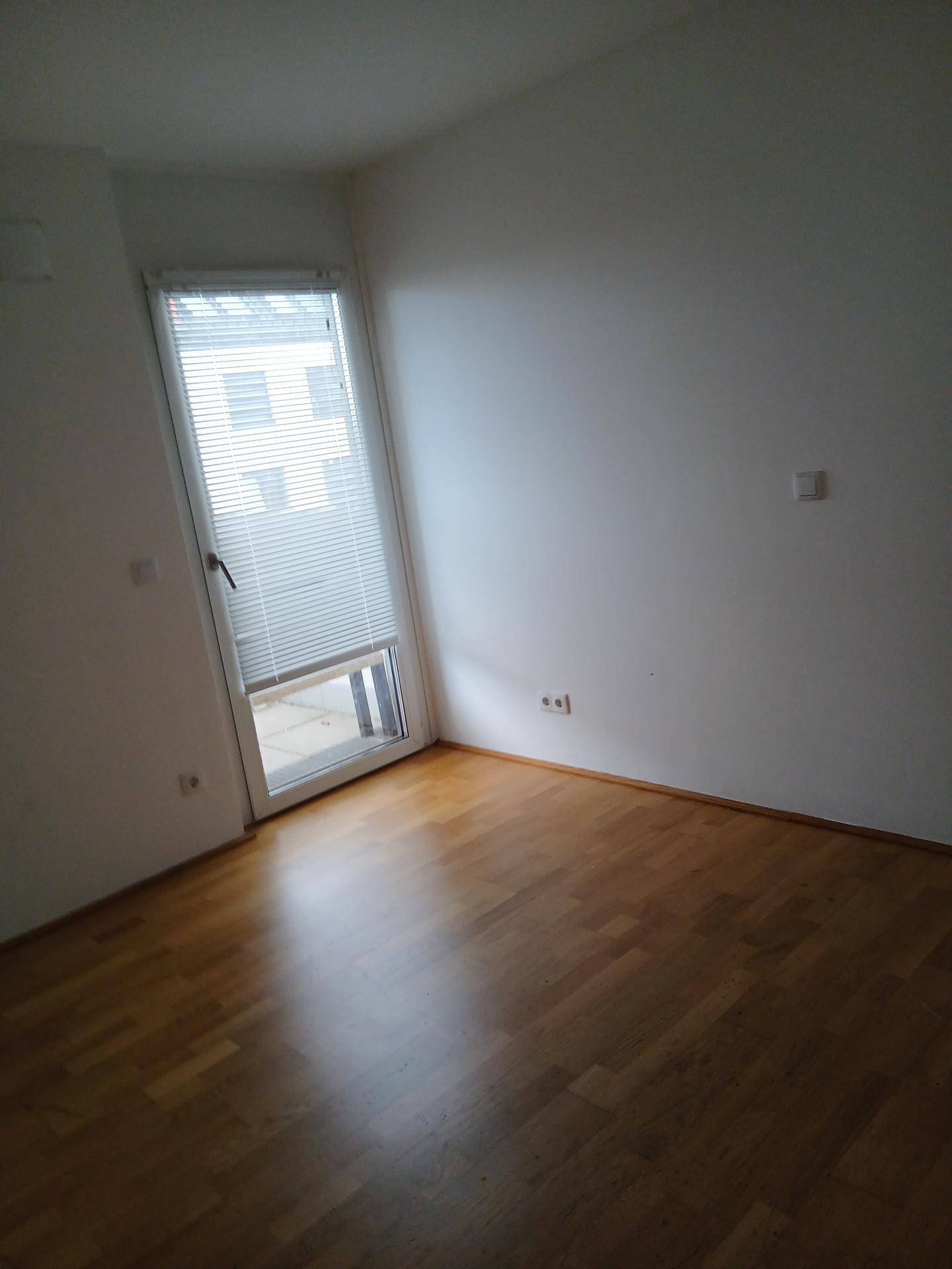 Leeres Zimmer mit Holzboden und einer Tür, die Zugang zu einem Balkon bietet.