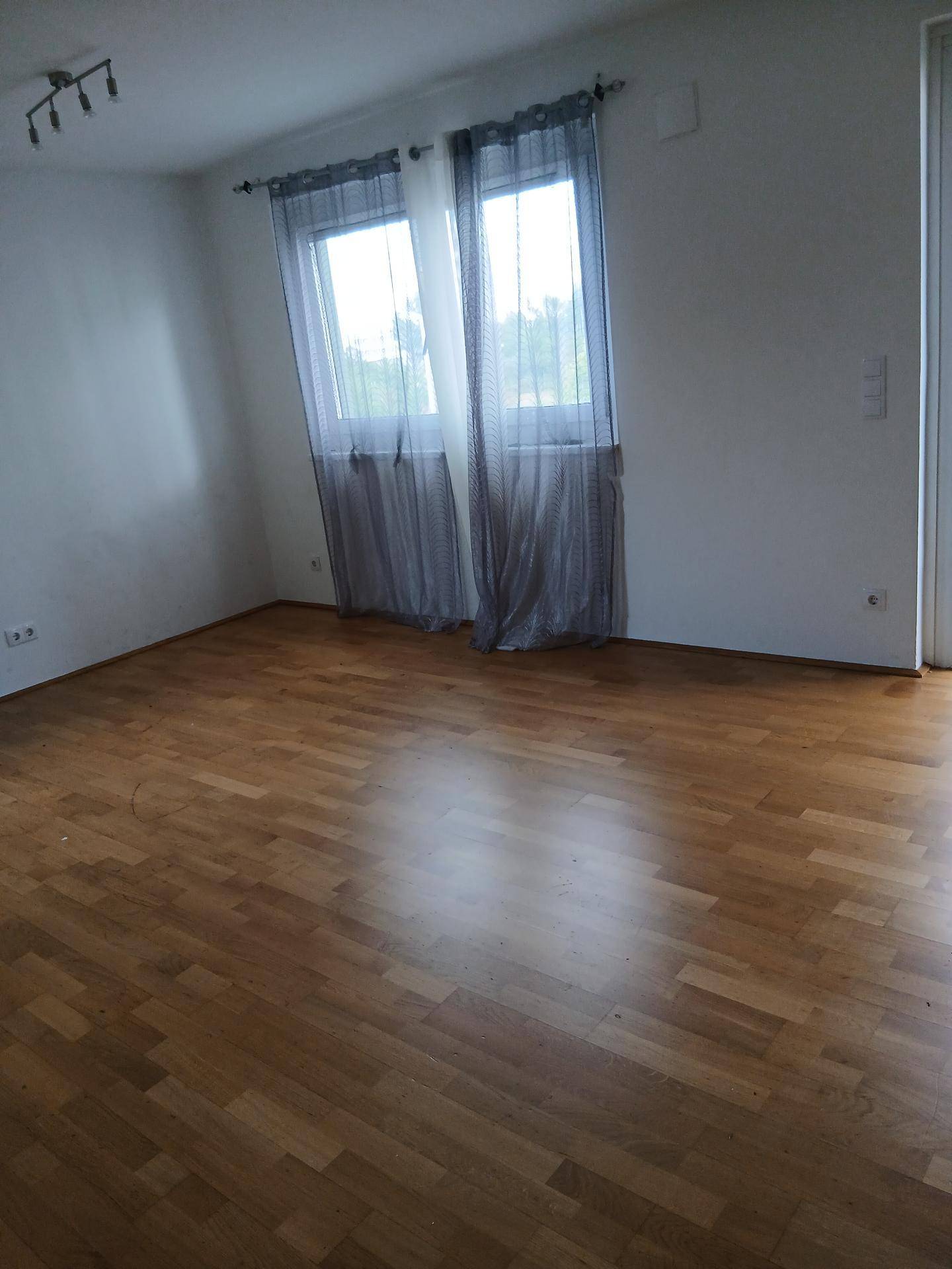Helles Zimmer mit Holzboden und zwei Fenstern, die viel Licht hereinlassen.