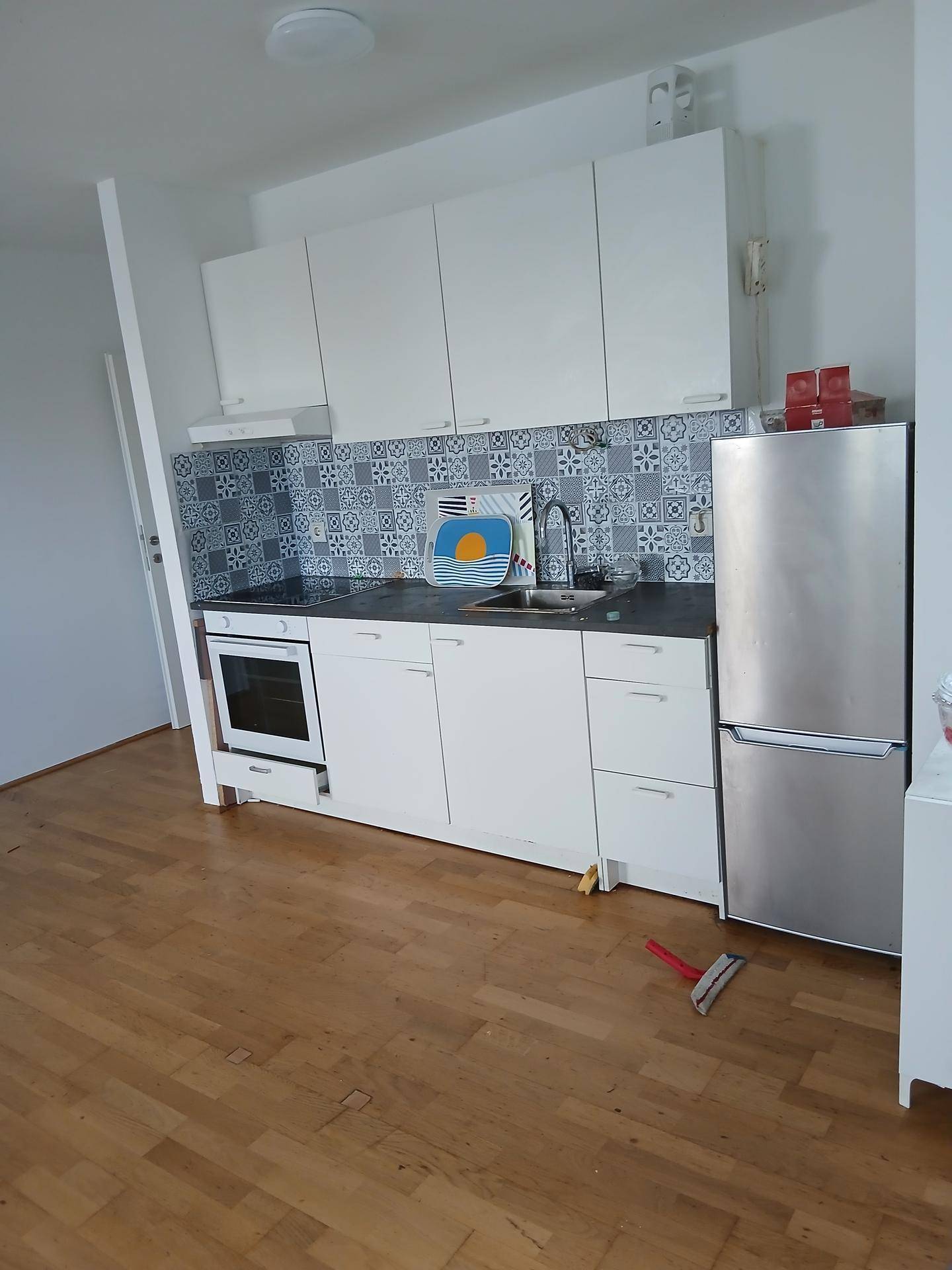 Kleine Einbauküche mit weißen Schränken, Herd und Kühlschrank, offen zum Wohnbereich.