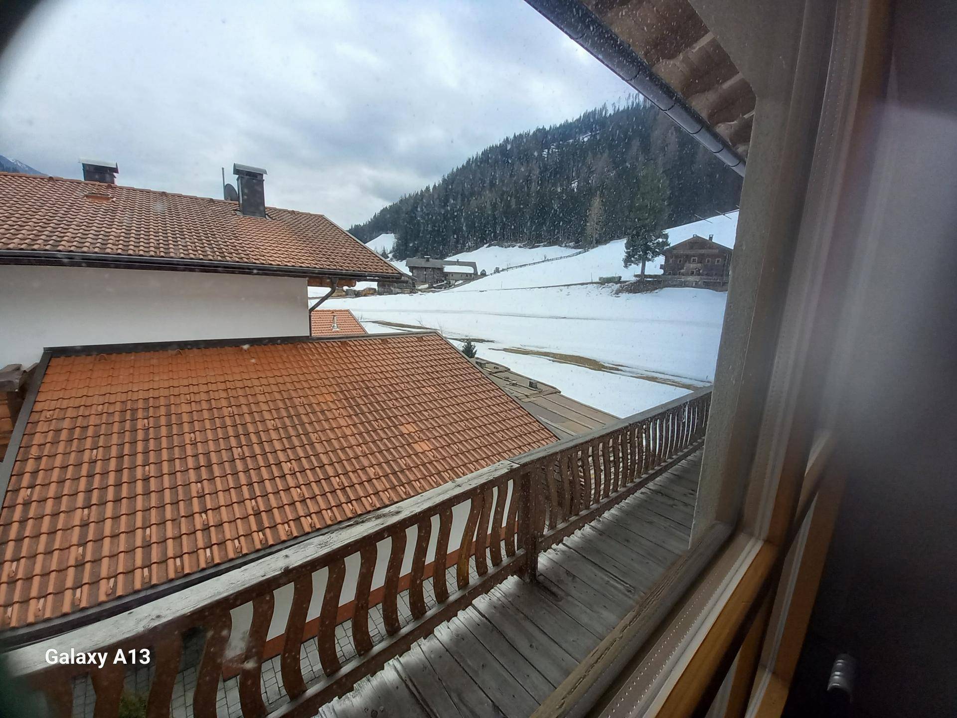 Blick aus dem Fenster auf einen Balkon mit Holzgeländer und die verschneite Berglandschaft.