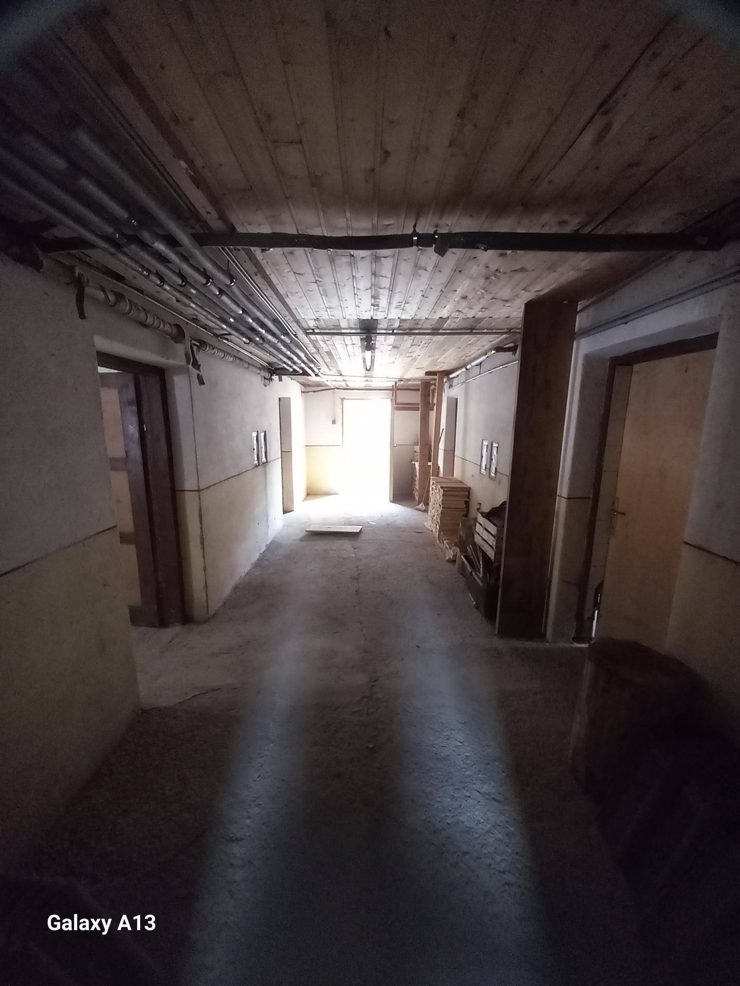 Langer Flur im Untergeschoss mit Betonboden und sichtbaren Rohren an der Holzdecke.