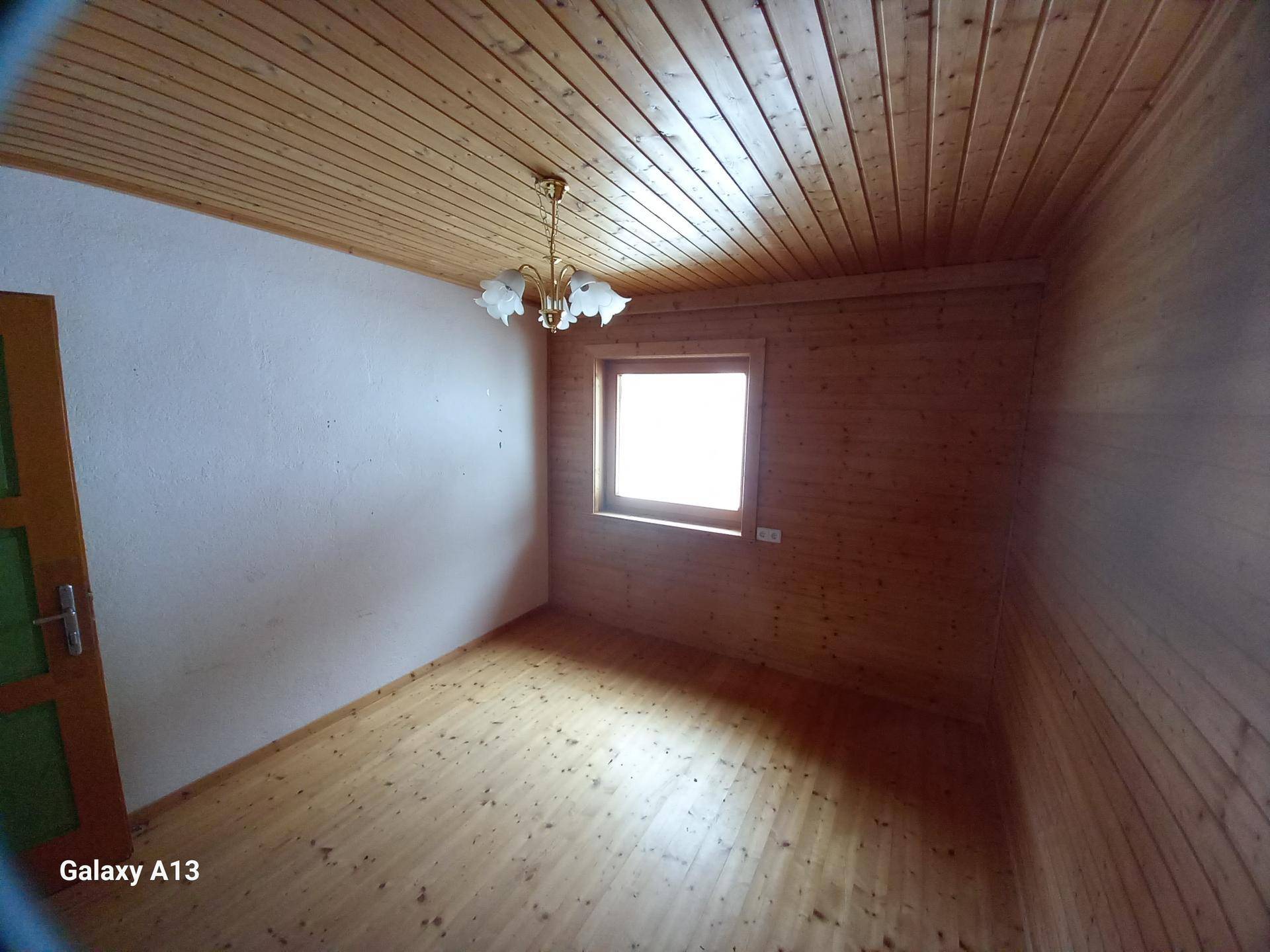 Leeres Zimmer mit Holzwänden, Holzdecke und Holzboden, beleuchtet durch ein Fenster.