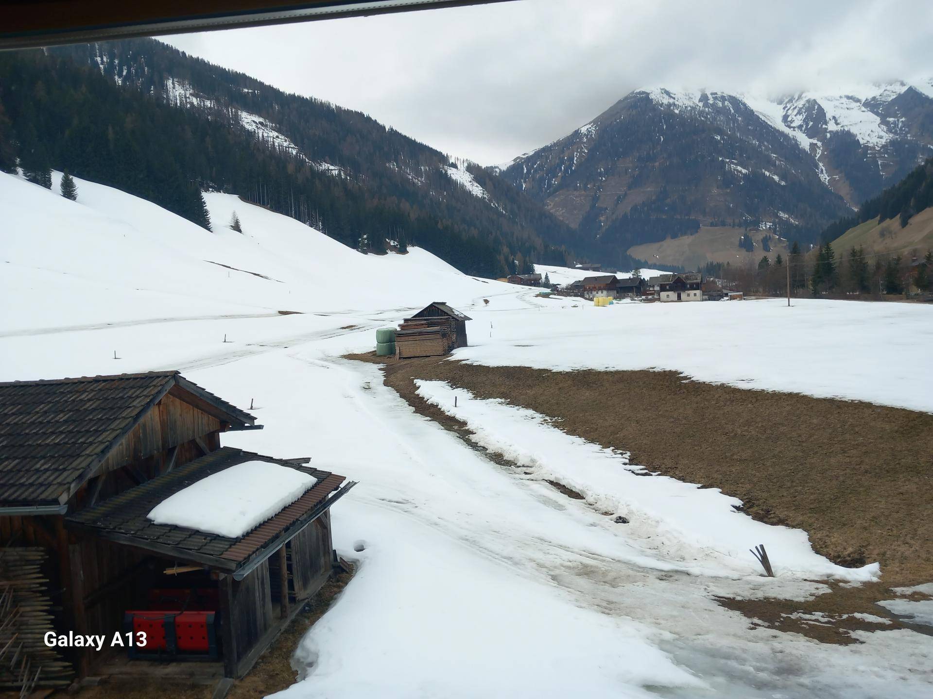 Weitläufiger Blick auf eine verschneite Tallandschaft mit vereinzelten Gebäuden und Bergen im Hintergrund.