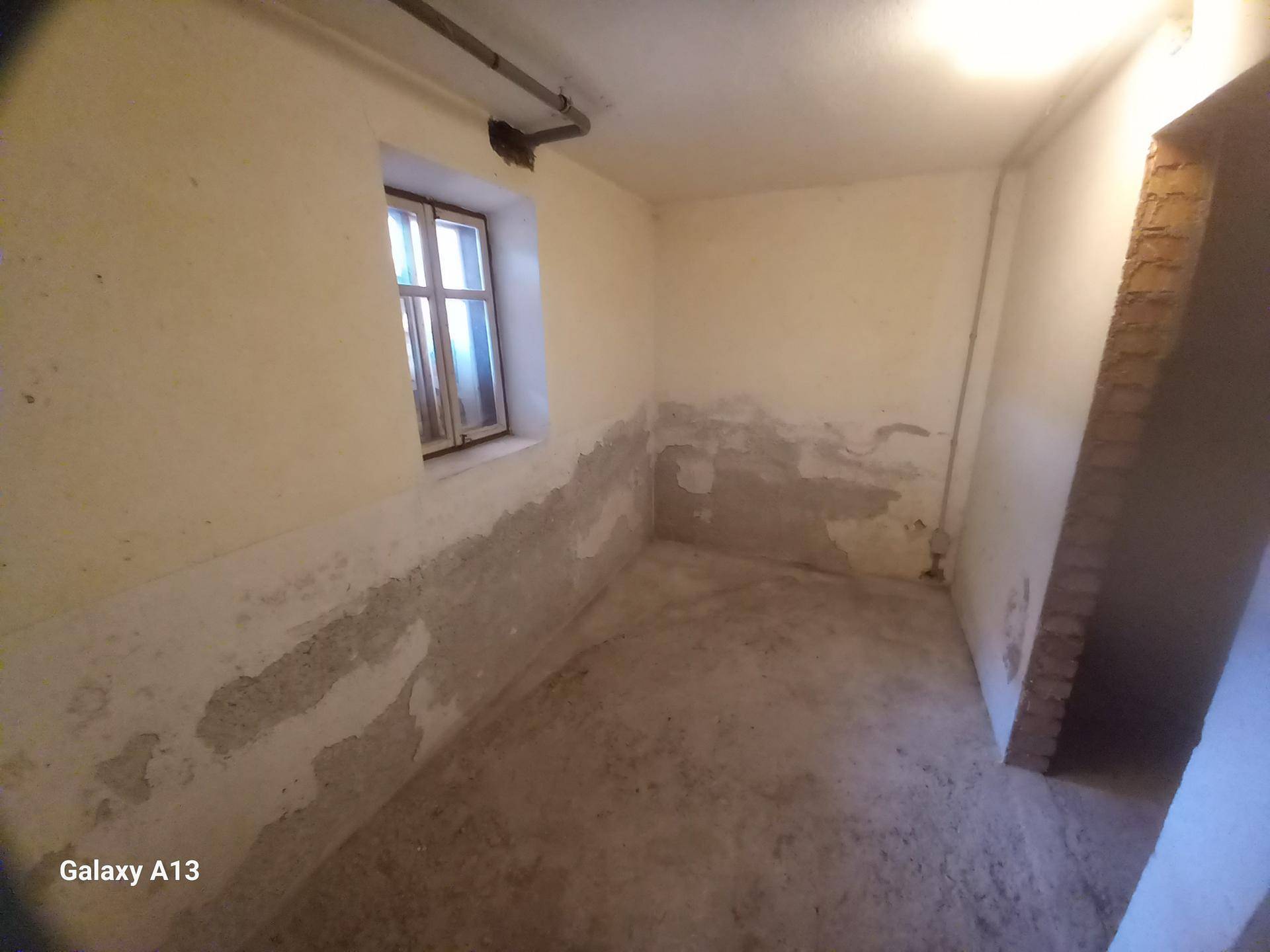 Kleiner Raum im Untergeschoss mit Betonboden, einem Fenster und abgenutzten Wänden.