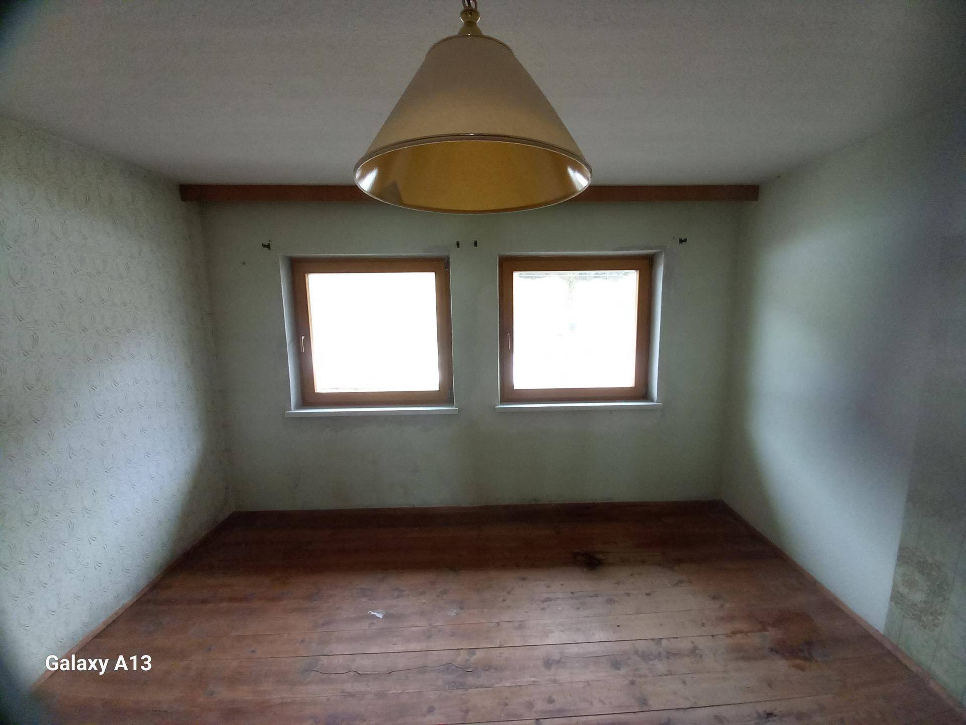 Leeres Zimmer mit zwei Fenstern, Holzboden und einer einfachen Deckenleuchte.