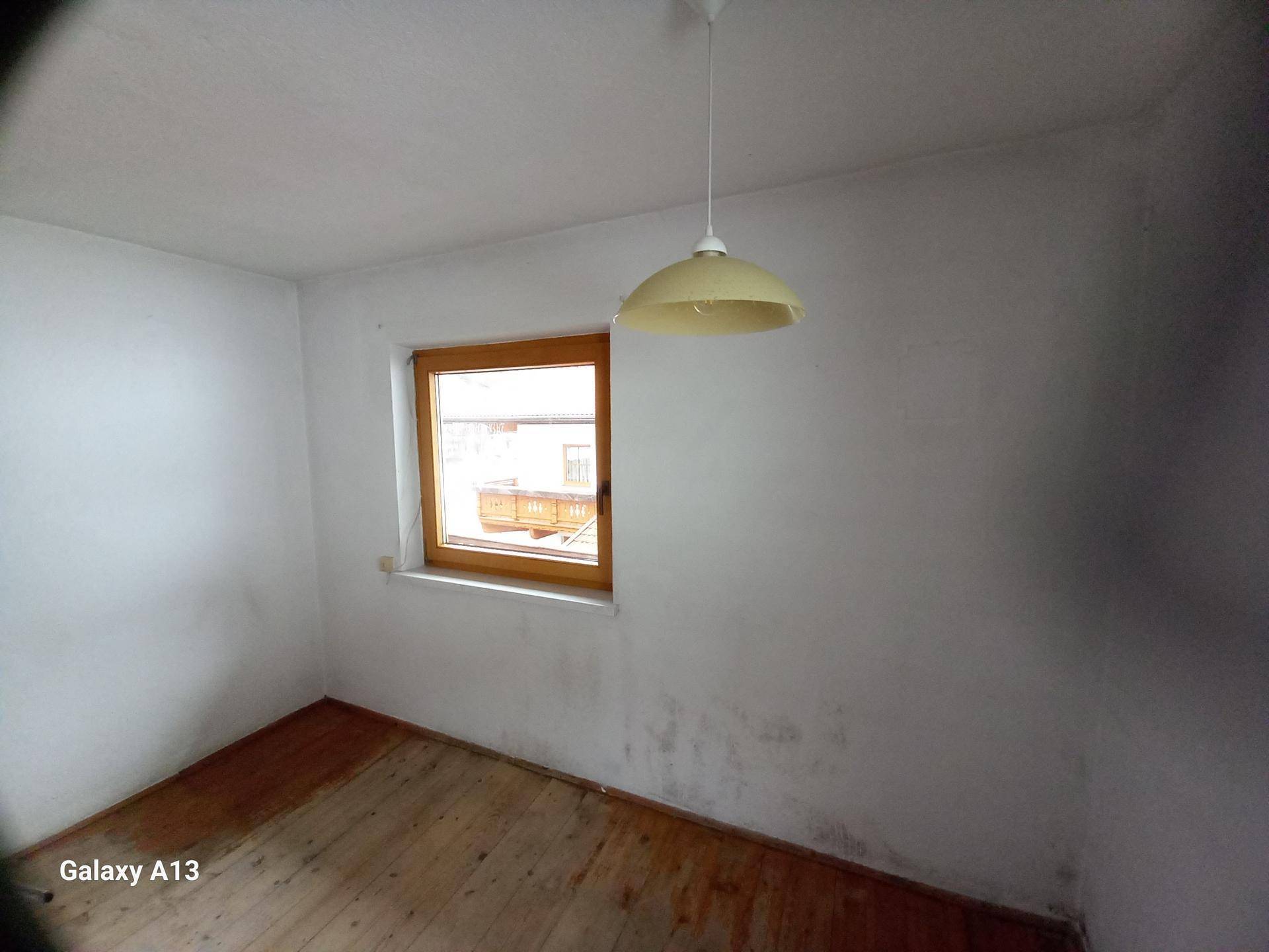 Helles Zimmer mit einem Fenster, Holzboden und weißen Wänden, die leichte Abnutzungsspuren zeigen.