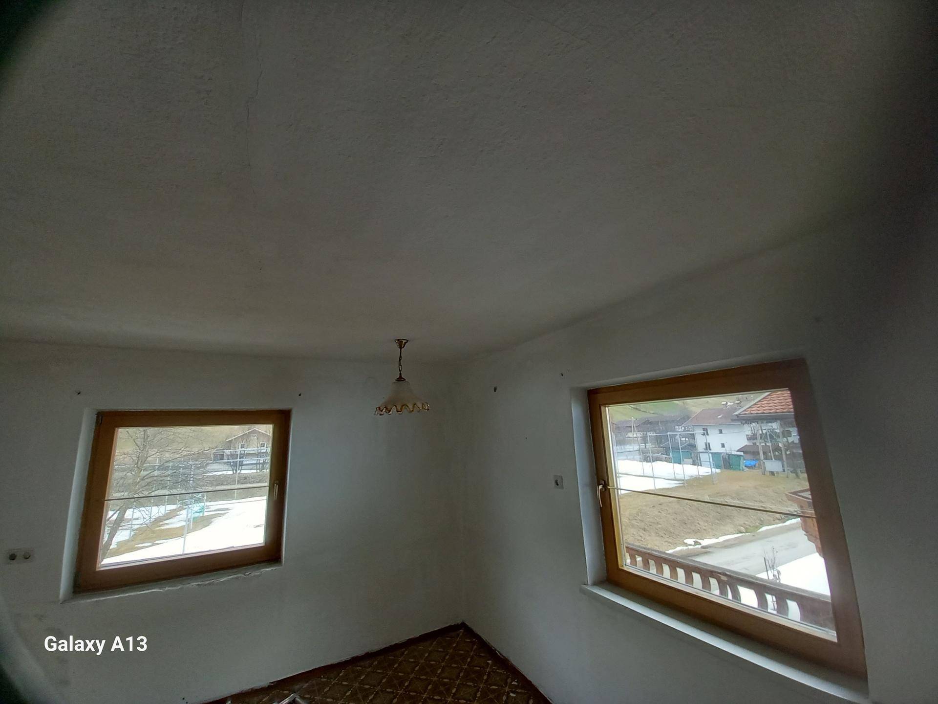 Leeres Zimmer mit zwei Fenstern, gemustertem Boden und einer einfachen Deckenleuchte.