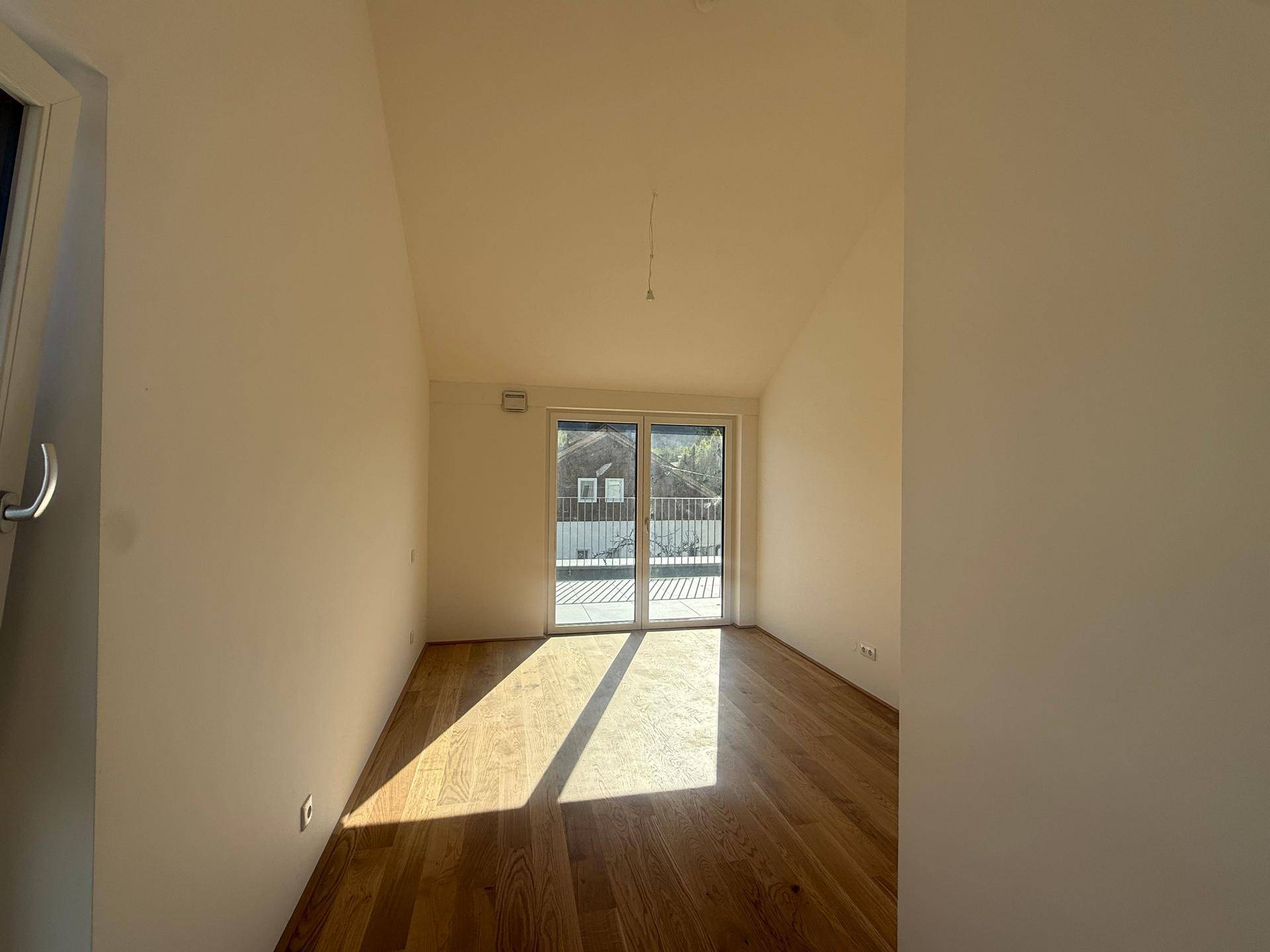 Helles Schlafzimmer mit Holzboden, schrägen Decken und großem Schiebetürfenster zum Balkon.